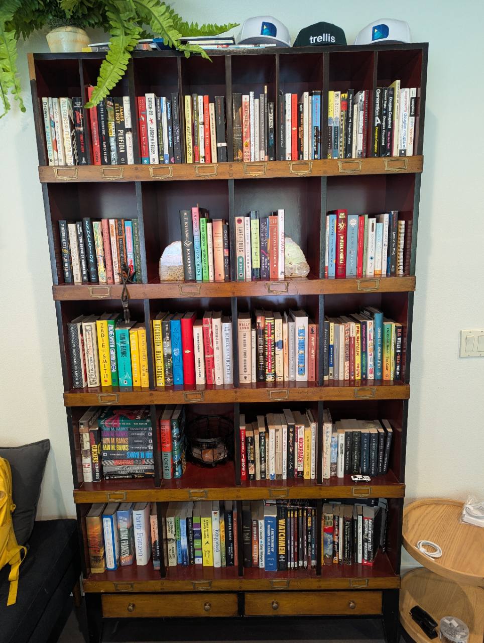 Original photograph of the dark wood bookshelf with five rows of books, brass label holders, a fern and caps.